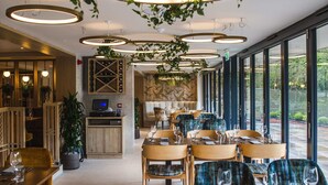Un restaurante con mesas redondas, sillas y plantas colgantes. Hay una barra y una zona de estar con vistas al exterior.