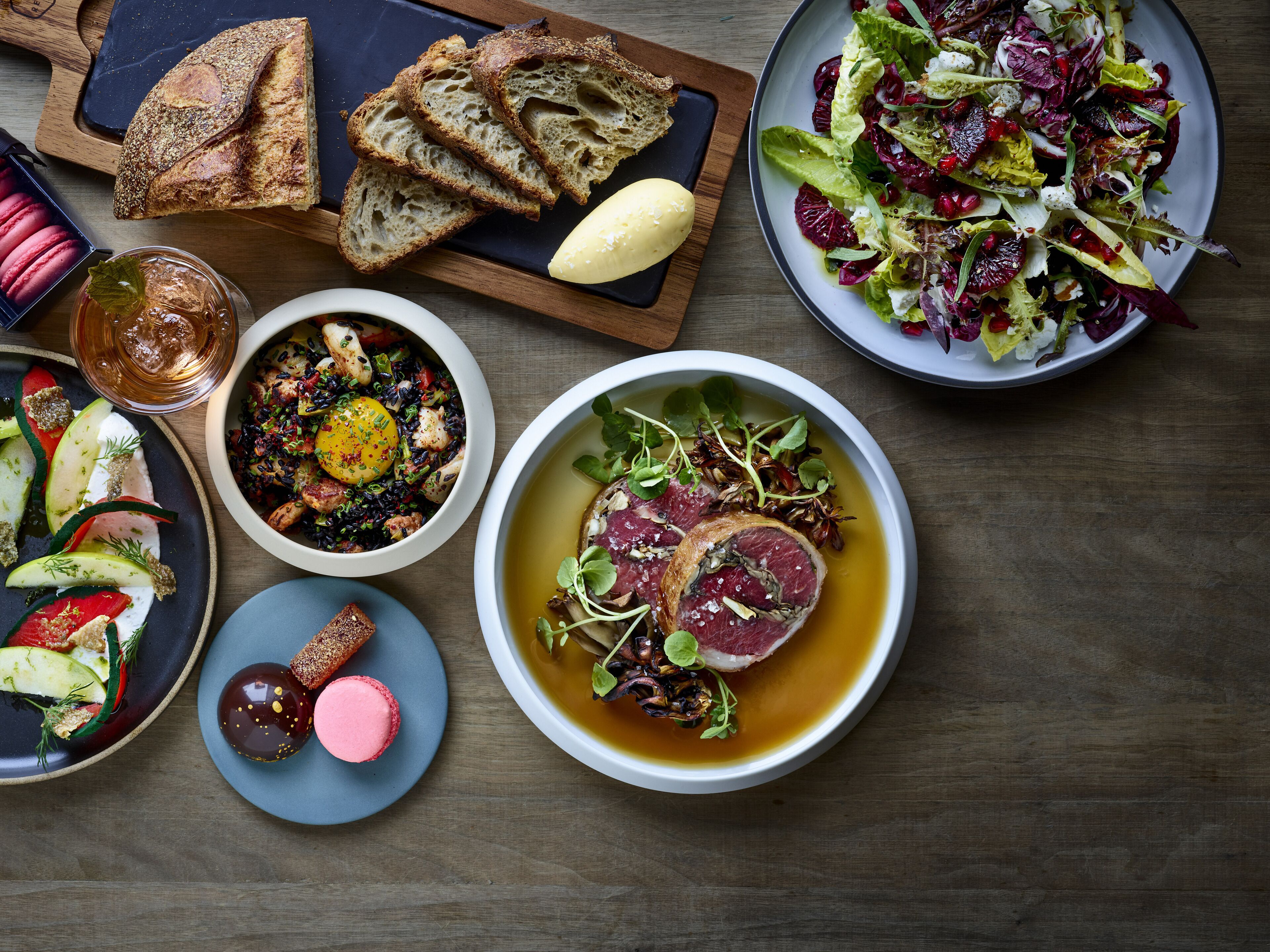 A variety of dishes including a salad, soup, and a plate with meat, along with slices of bread and a glass of drink on a wooden table.