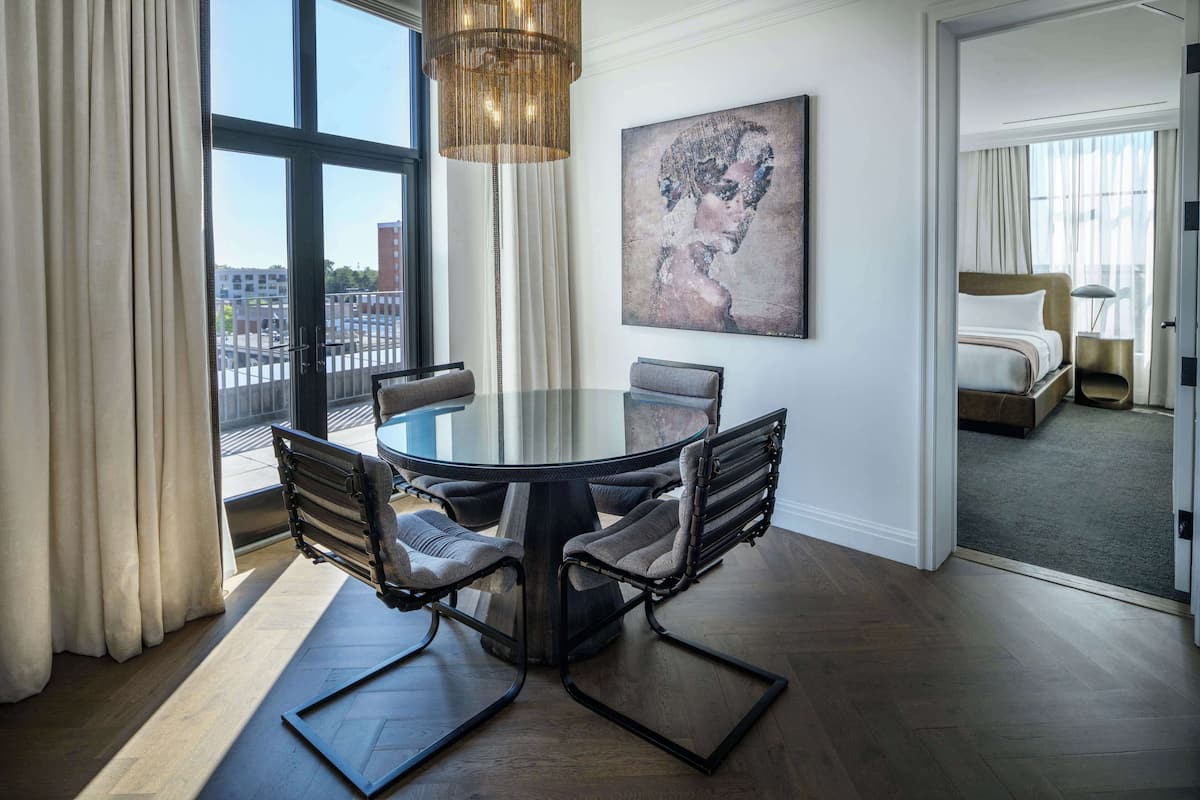 Ett modernt hotellrum med ett runt matbord, ett stort fönster med gardiner, en tavla på väggen och utsikt över staden.