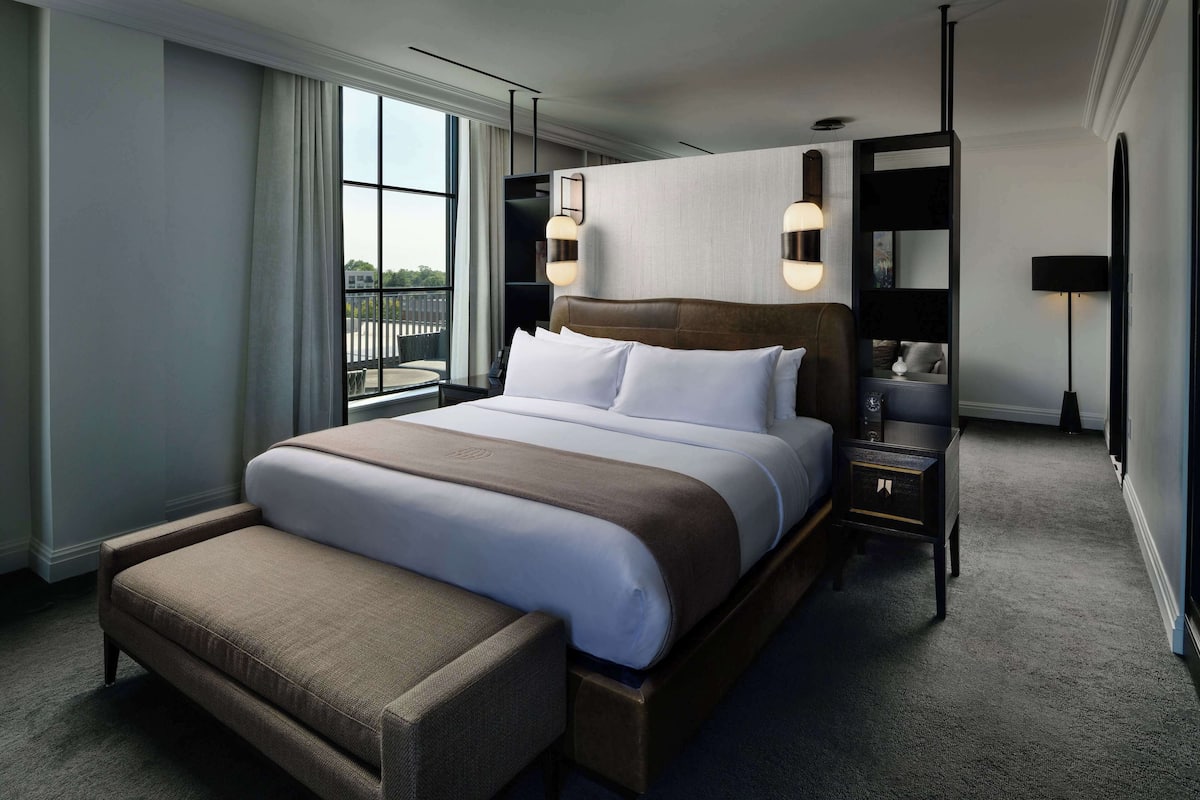 Ett modernt hotellrum med en stor säng, ett sängbord, en liten avlastningsyta och utsikt över omgivningarna genom ett fönster.