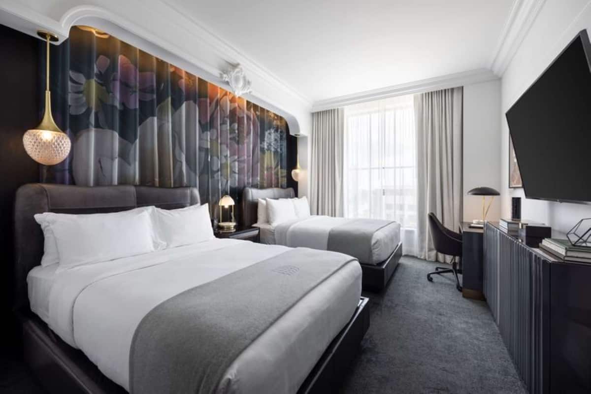 Ett hotellrum med två sängar, ett skrivbord, en stol, en tv och ett stort fönster med gardiner.