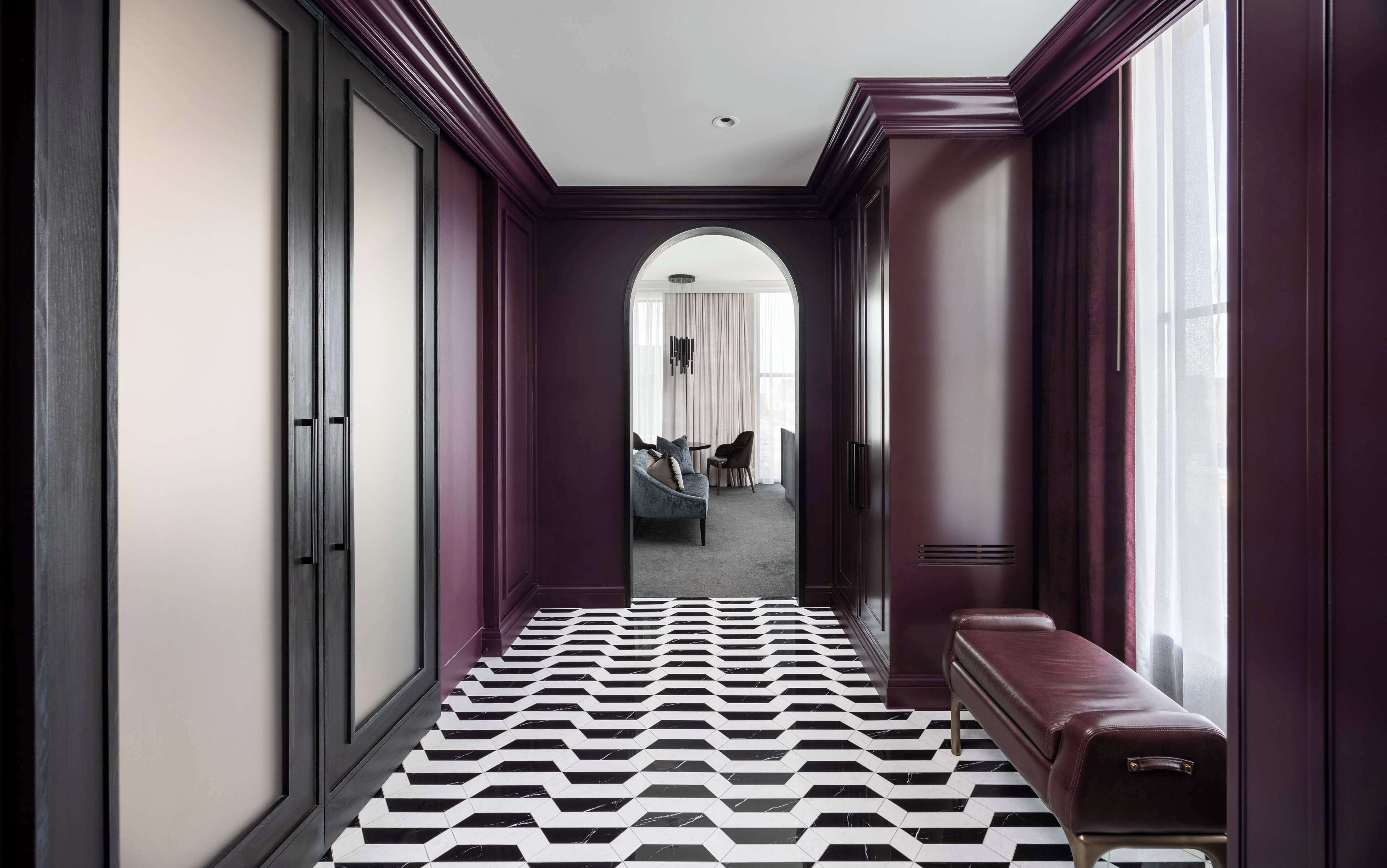 A hallway with a black and white patterned floor, a bench, and a view into a room with a bed and curtains.