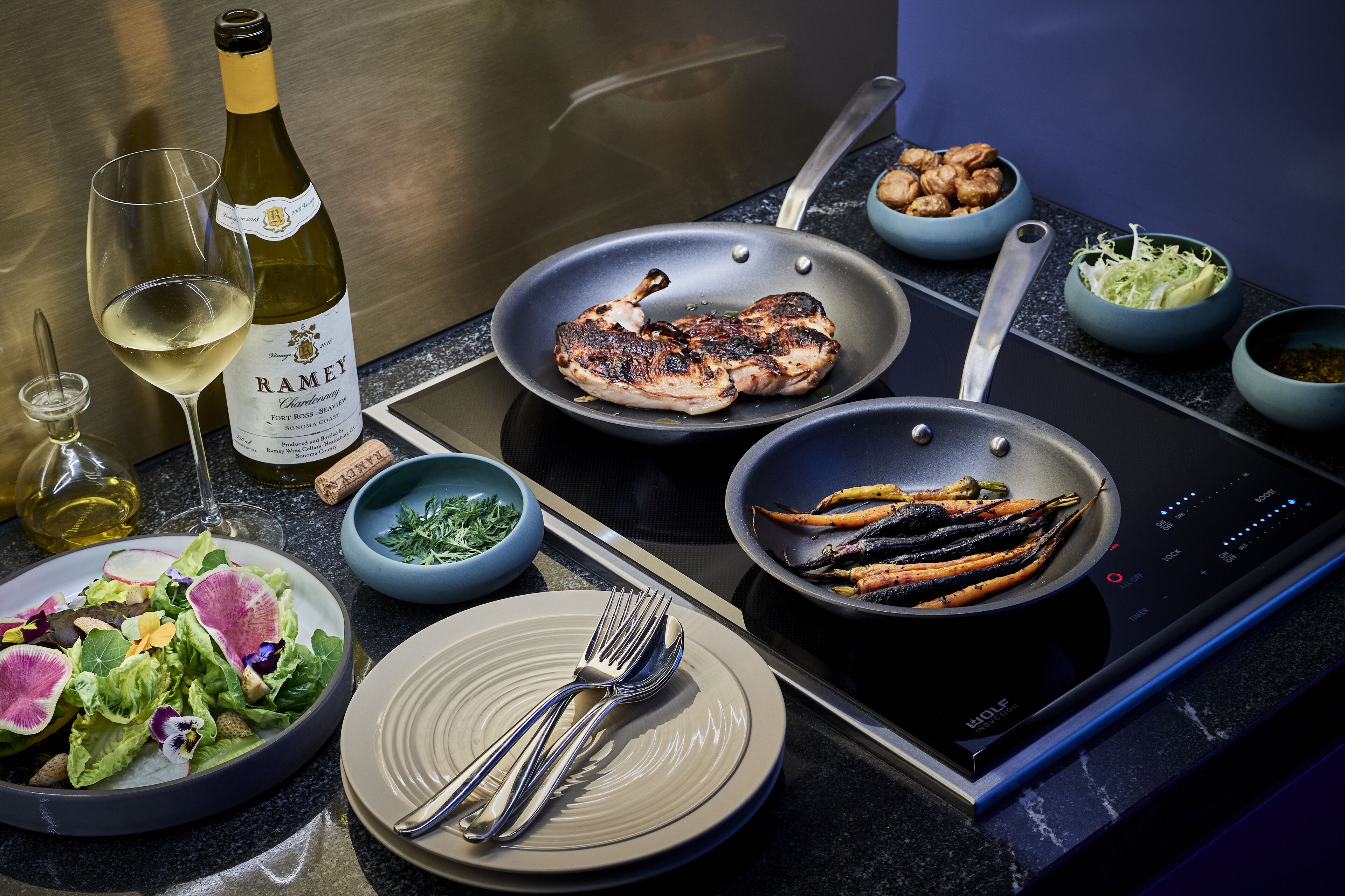 A kitchen counter with a pan of grilled chicken, a bottle of wine, and various bowls of food.