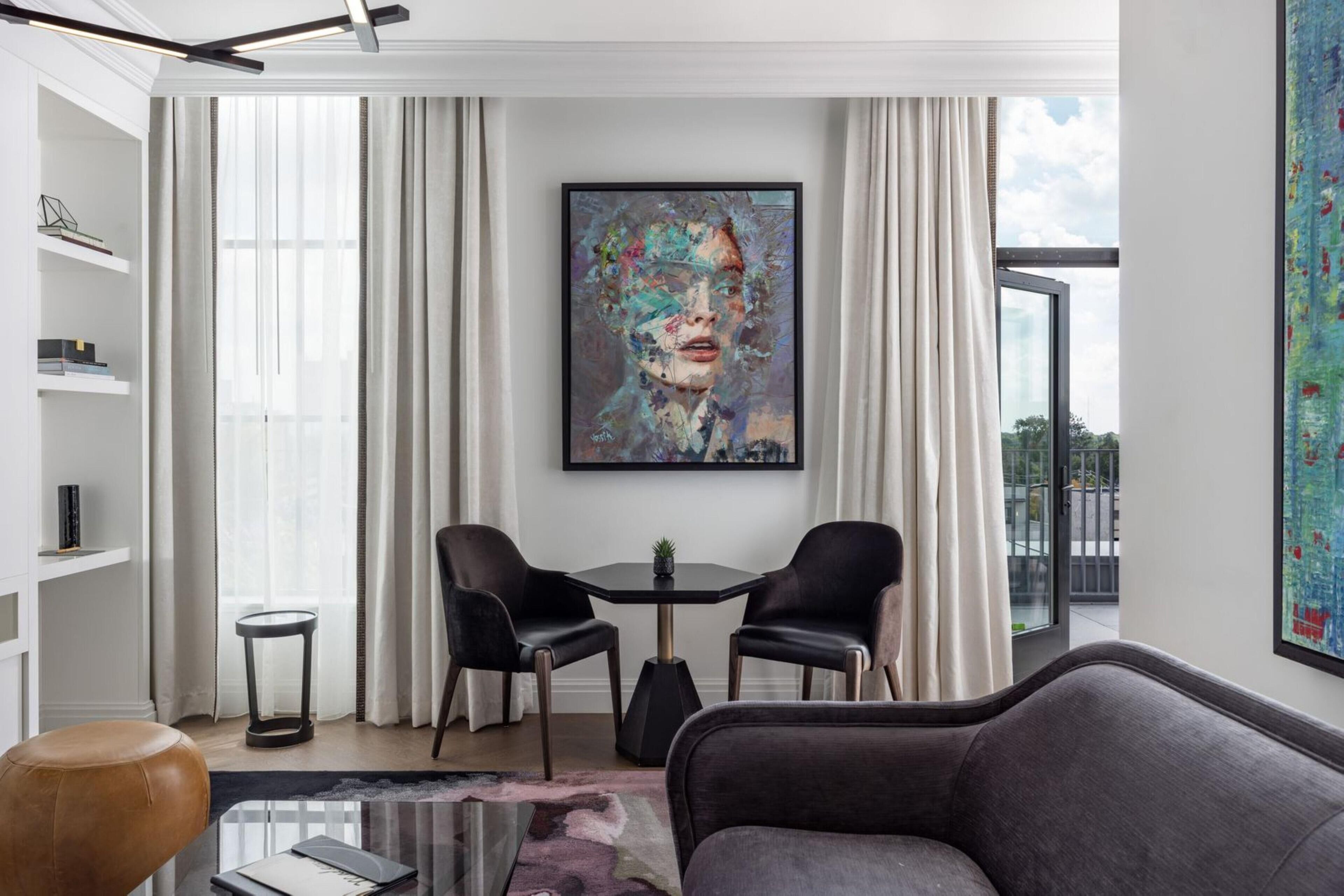 A modern living room with a large abstract painting, two black chairs, a round table, and a glass coffee table.