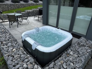 A hot tub surrounded by a stone border, with a patio table and chairs nearby.