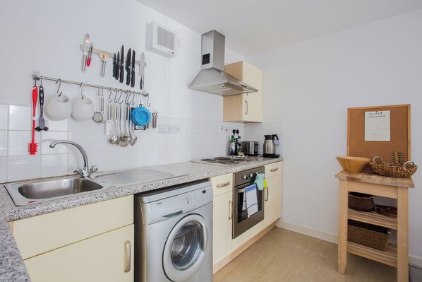 A modern kitchen with a built-in dishwasher, a stove, and a microwave. There is a hanging rack with kitchen utensils and a corkboard with a welcome sign.
