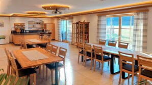 A spacious dining area with wooden tables and chairs, a kitchen in the background, and large windows allowing natural light. - Ferienwohnung Morgensonne, 60 qm Obergeschoss, Separates Schlafzimmer (Söchtenau)