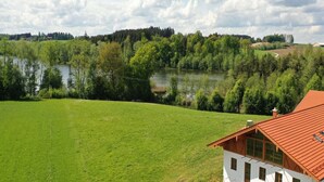 Un edificio blanco con techo rojo, rodeado por un campo verde exuberante y un lago, con árboles y una colina al fondo.