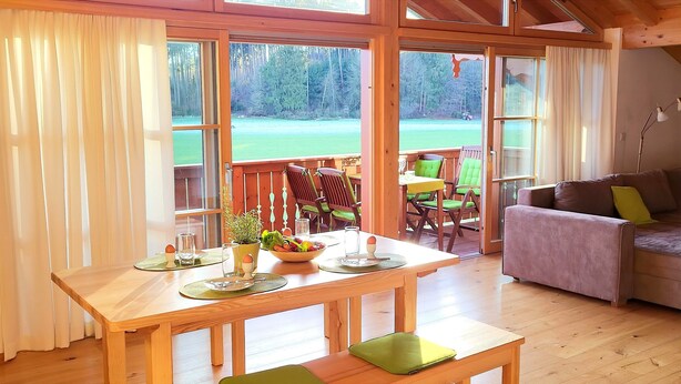 Habitación con piso de madera, una mesa de comedor para dos personas, un sofá y vista a un área exterior con césped a través de ventanas amplias.