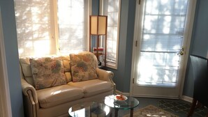 Un salon avec un canapé beige, deux tables en verre, un tapis à motifs et une porte avec des stores.