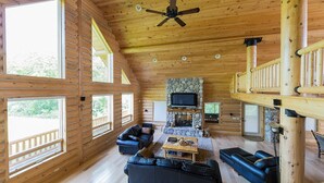 L’intérieur d’une cabane en rondins, avec une cheminée en pierre, des canapés en cuir, un téléviseur et un balcon.