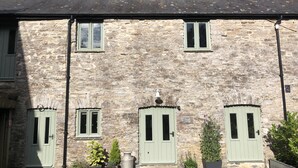 A stone building with three green doors and windows, a small garden area with potted plants, and a sign indicating "Cottage." - Two bedroom stone cottage near Dartmoor and Devon beaches (Ashburton)