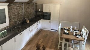 A kitchen with white cabinets, a stone wall, a wooden ceiling, and a dining area with white chairs and a table set for four. - Two bedroom stone cottage near Dartmoor and Devon beaches (Ashburton)