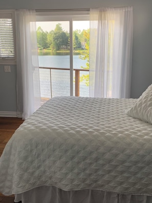 Slaapkamer met een groot bed, uitzicht op een meer en een raam met dunne gordijnen.