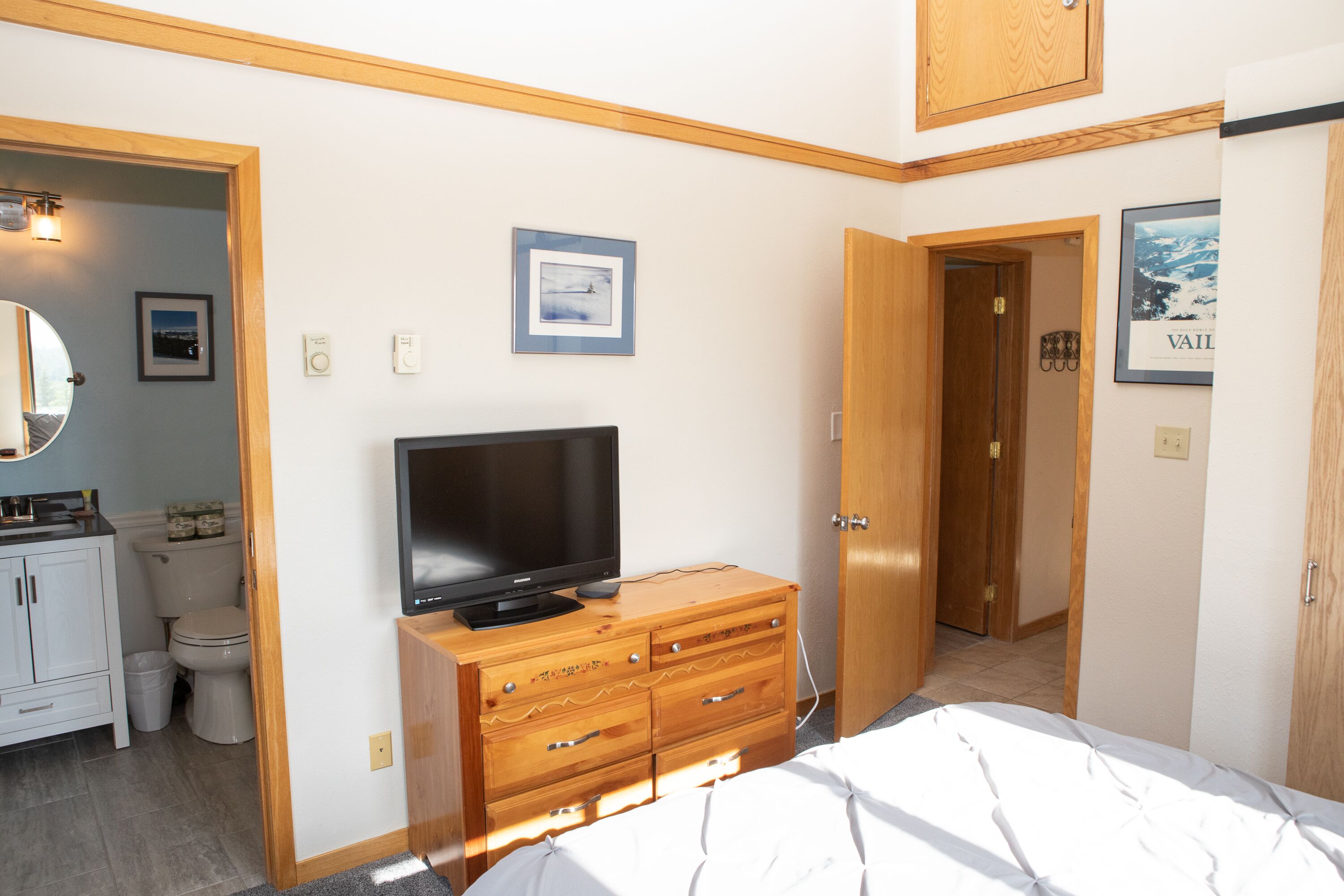 Une chambre à coucher avec un lit, une commode en bois, un téléviseur et une salle de bain visible à travers une porte ouverte.