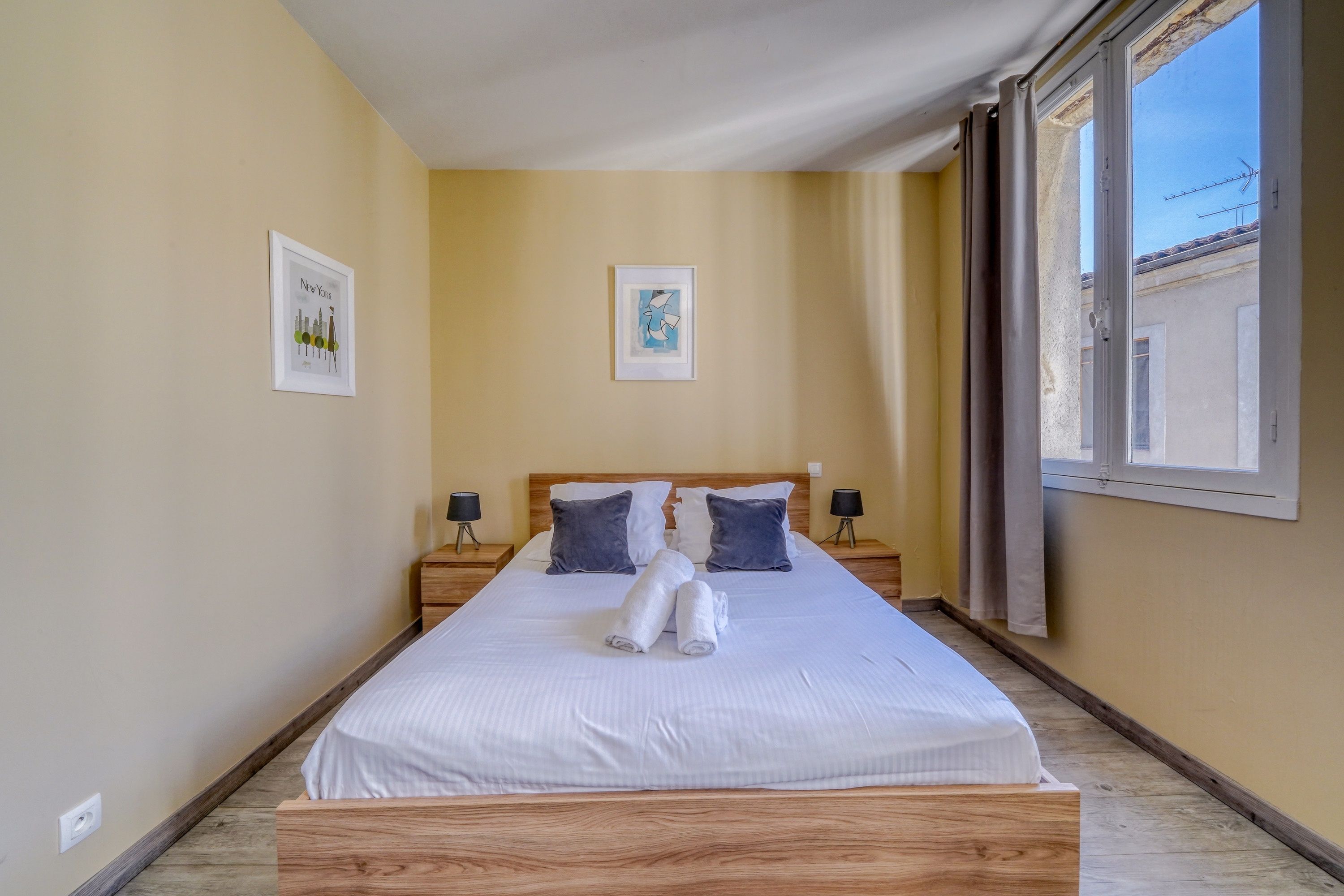 A hotel room with a bed, two bedside tables, a window with curtains, and two framed pictures on the wall.