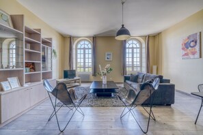 A spacious living room with a black sofa, two leather chairs, a coffee table, and a TV. There are large windows with curtains, a bookshelf, and a painting on the wall. - LE RICHELIEU - Magnifique duplex avec une vue Incroyable (Bordeaux)