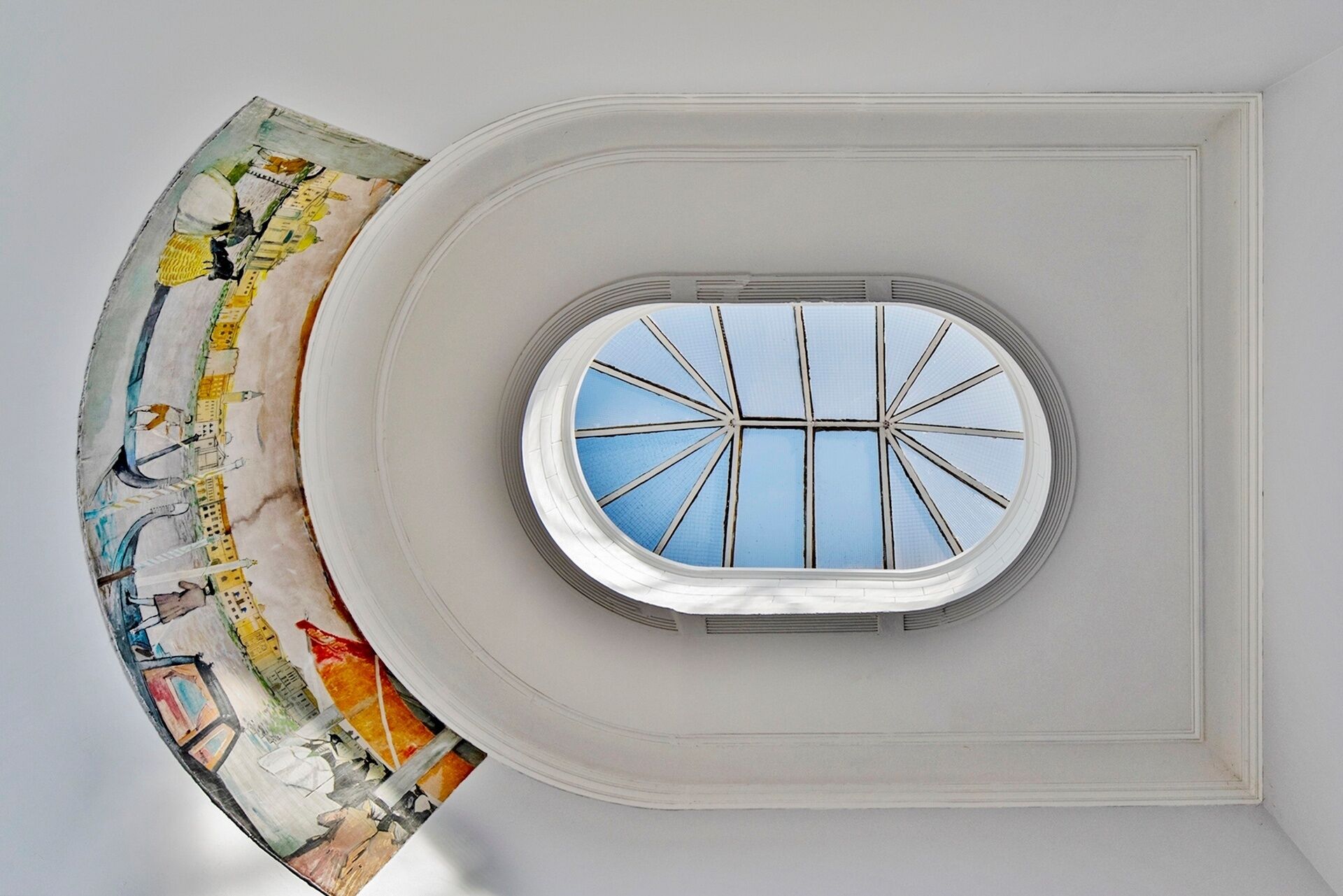 A circular skylight with a grid pattern, a white ceiling, and a colorful mural on the adjacent wall.