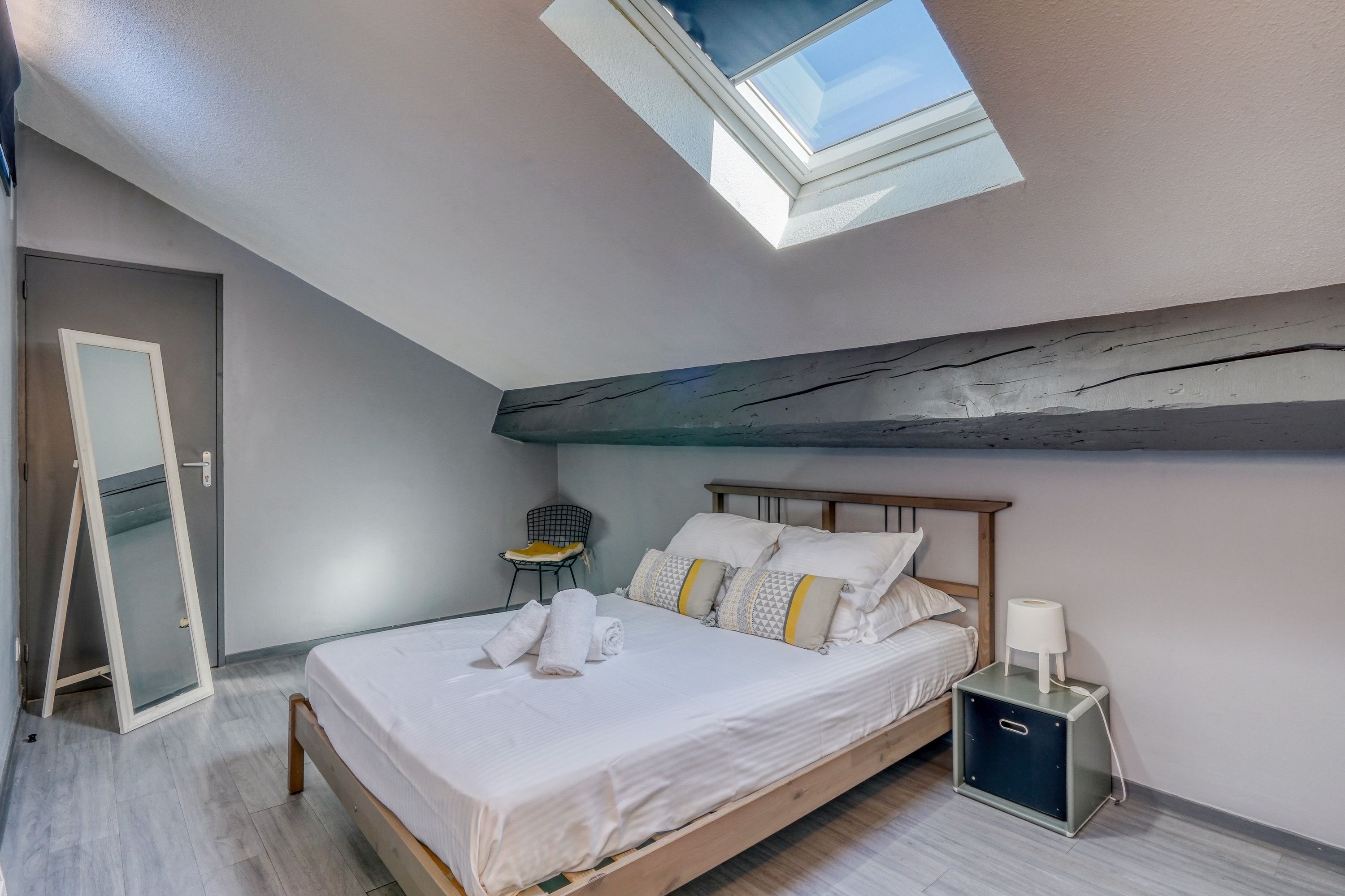 A bedroom with a skylight, a bed with pillows, a chair, and a bedside table with a lamp.