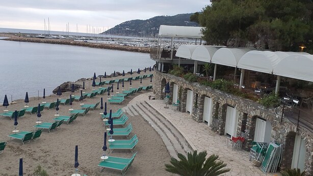Una spiaggia con file di sedie a sdraio verdi e ombrelloni blu, un edificio in pietra con archi e, sullo sfondo, barche e un ponte.