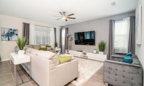 Ein modernes Wohnzimmer mit einem beigen Sofa, einem grauen Sessel, einem Flachbildfernseher und einem Deckenventilator.