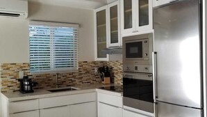 A modern kitchen with white cabinetry, a stainless steel refrigerator, a built-in oven, and a window with blinds.