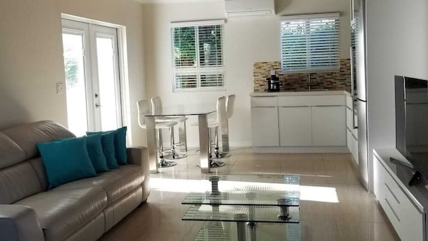 A modern living room with a glass coffee table, a sofa with teal cushions, and a kitchen area with white cabinets and a built-in oven.