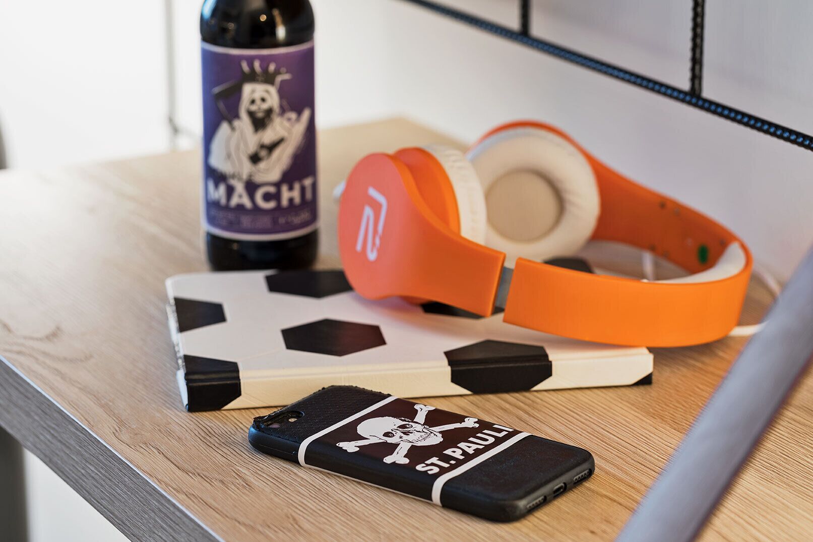 A smartphone with a skull and crossbones case, a bottle of drink, and orange headphones on a wooden surface.
