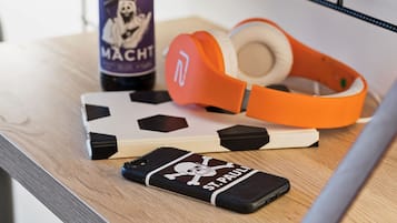 A smartphone with a skull and crossbones case, a bottle of drink, and orange headphones on a wooden surface.