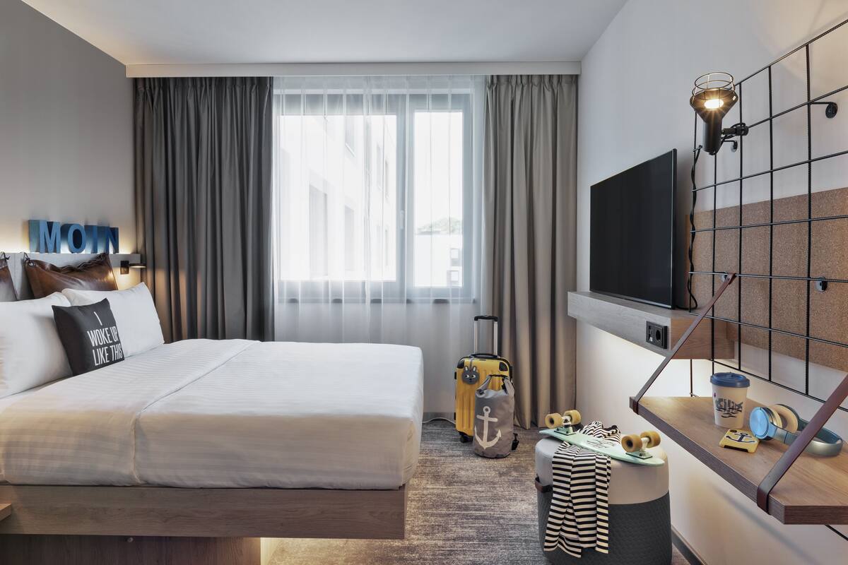 A hotel room with a large bed, a TV mounted on the wall, a desk with a suitcase, and a window with curtains.