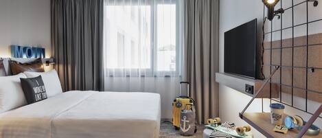 Habitación de hotel con una cama grande, un televisor montado en la pared, un escritorio con una maleta y una ventana con cortinas.