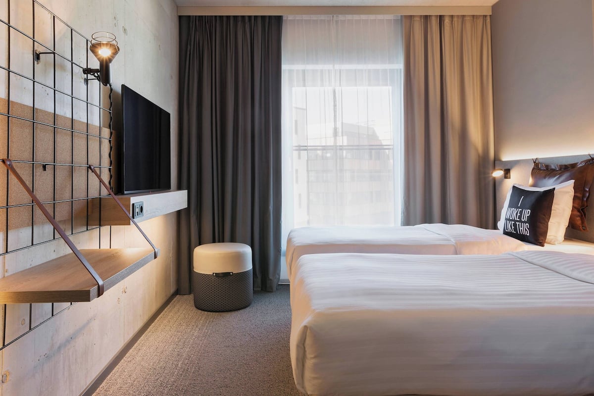 A hotel room with a TV mounted on a wall, a bed, a small ottoman, and a window with curtains.