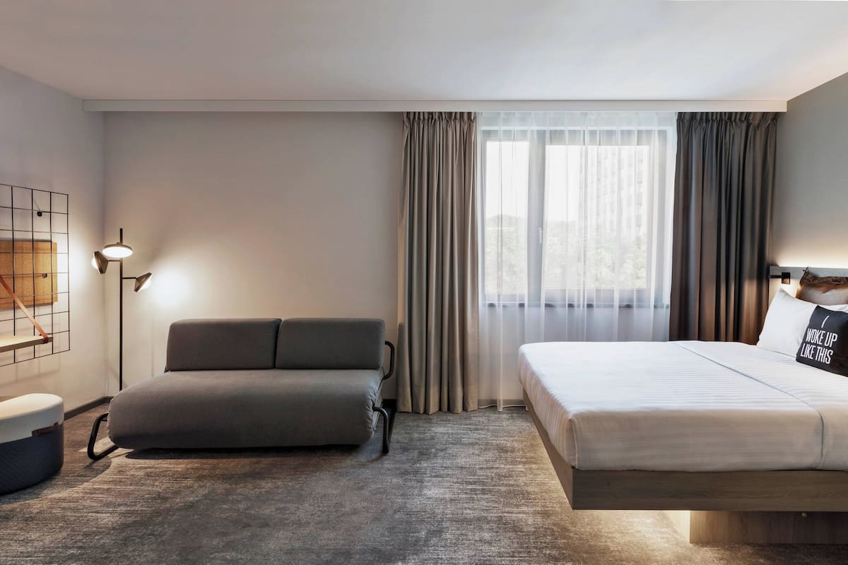 A modern hotel room with a sofa, a bed, a floor lamp, and a view of the outdoors through sheer curtains.