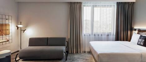 Una habitación de hotel moderna con sofá, cama, lámpara de pie y vistas al exterior a través de cortinas translúcidas.