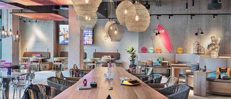 Een modern café met een lange houten tafel, rieten stoelen en hangende rotanlampen.