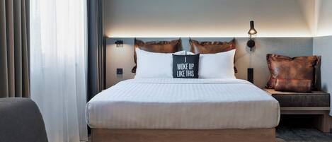 Ein modernes Hotelzimmer mit einem großen Bett, einem Nachttisch, einer Bank und Blick auf die Stadt durch transparente Vorhänge.