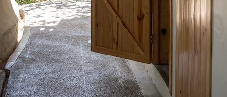 A wooden door with a diagonal brace design, leading to a covered outdoor area with a concrete floor and a view of greenery.