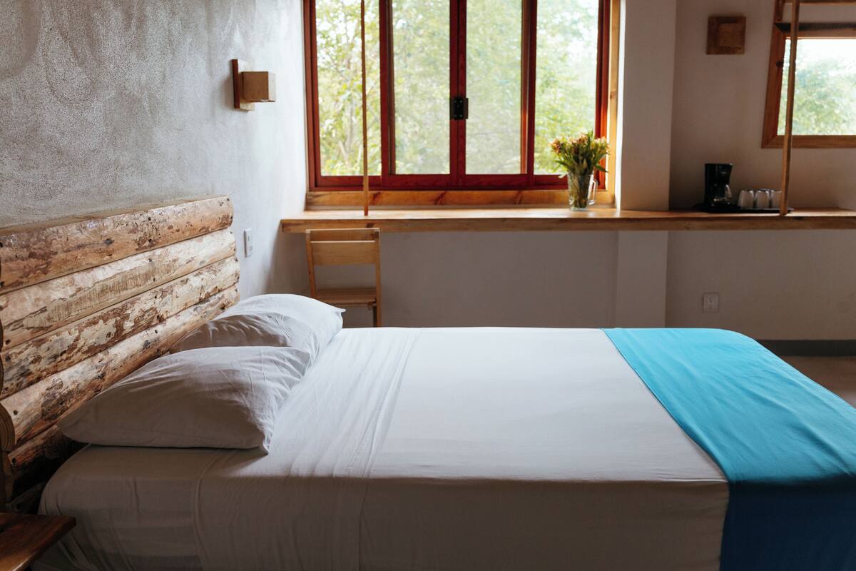 A bedroom with a wooden headboard, a bed with white bedding, a window with a view of trees, and a small table with a vase of flowers.