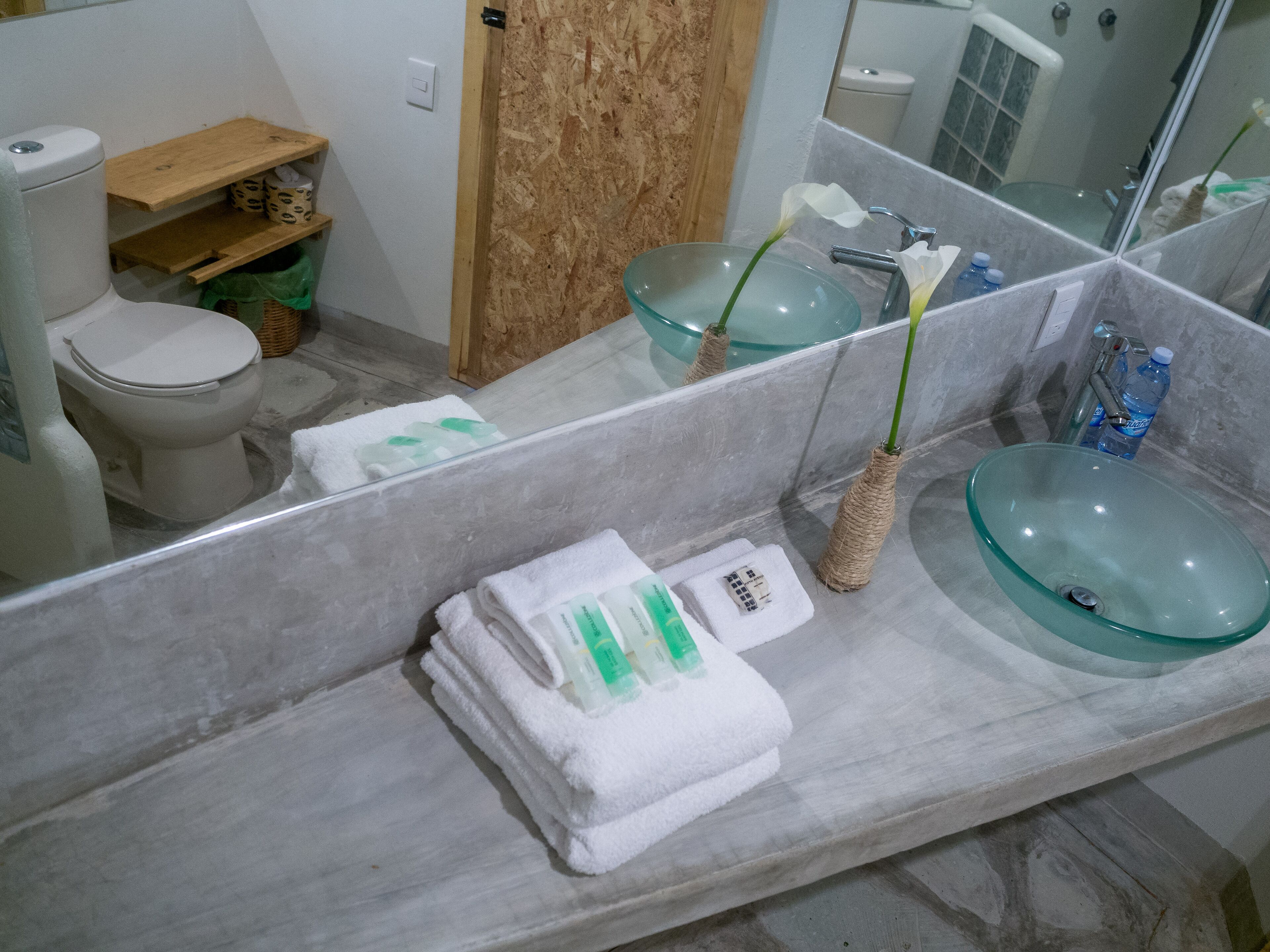Un baño con un lavamanos de vidrio, un inodoro, un estante de madera y un espejo.