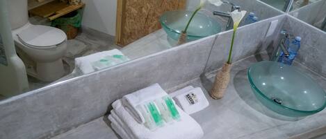 Un baño con un lavamanos de vidrio, un inodoro, un estante de madera y un espejo.