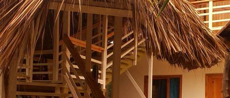 A wooden staircase leading to a thatched-roof structure, a wooden door, and a palm tree.