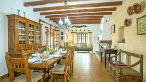 A dining area with a wooden table set for four, flanked by wooden chairs with cushioned seats. A wooden cabinet with glass doors is to the left, and a bench with a cushion is to the right. The room has a tiled floor and wooden beams on the ceiling. - 3 Bedrooms, 2 bathrooms, enbeded into the natural mediterranean surrounding. (Pollença)