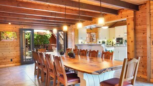 A wooden dining area with a large table and chairs, a kitchen in the background, and a view of the outdoors.