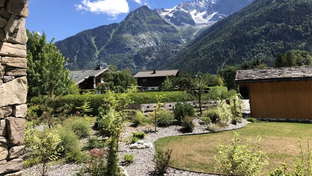 A mountain view with a stone building, a wooden house, and a garden with a gravel path.