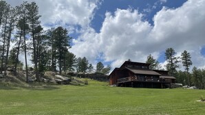 A wooden cabin with a balcony, surrounded by a grassy field and tall pine trees under a partly cloudy sky. - TRI-LEVEL HOUSE WITH HUGE DECK AND AMAZING VIEWS OF THE BLACK HILLS (Custer)