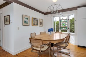 A dining room with a round wooden table, six wicker chairs, and a vase with blue flowers. There are three framed pictures on the wall, and a chandelier hangs from the ceiling.