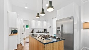 Une cuisine moderne avec des meubles blancs, un réfrigérateur en acier inoxydable, un îlot central doté d’une cuisinière à gaz et un éclairage suspendu.