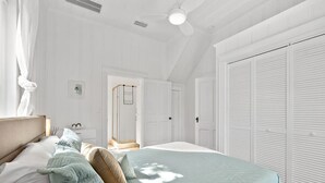 Une chambre avec un grand lit, des murs blancs, un ventilateur de plafond et une armoire.