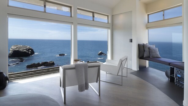 Ein modernes Zimmer mit großen Fenstern zum Meer, einem weißen Sofa und einer Bank mit Kissen.