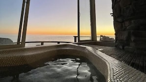 Ein Whirlpool mit Blick auf den Sonnenuntergang über dem Meer, umrahmt von einer Steinmauer und großen Fenstern.